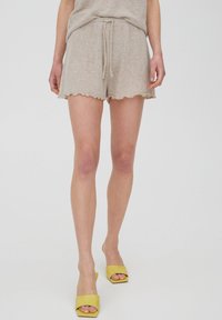 Beige shorts dengan tali serut dengan finishing tekstur, pinggiran bergelombang, dipasangkan dengan sepatu hak terbuka kuning di kaki telanjang. Desain yang sederhana dan santai.