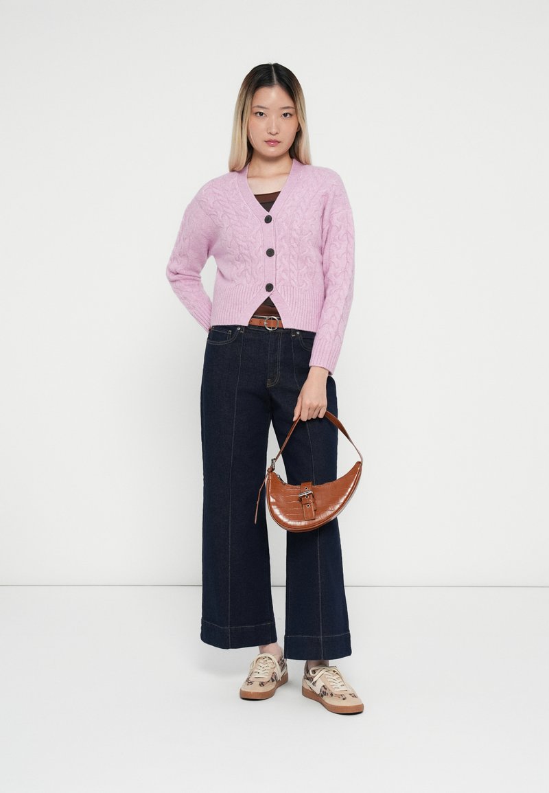 Cardigan en maille rose clair avec des boutons noirs, porté sur un haut marron. Un jean large foncé et un sac à main en cuir gaufré marron complètent le look.