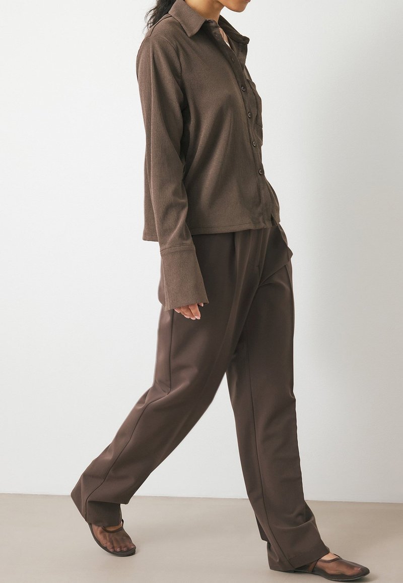 Brown long-sleeve button-up shirt features a textured fabric. Paired with loose brown trousers and transparent heeled sandals.