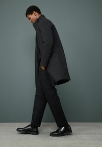 Abrigo negro largo con un cuello alto, llevado sobre un conjunto negro, combinado con zapatos de cuero negros. Textura suave, diseño minimalista.