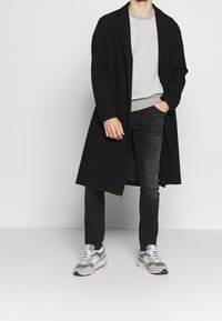 Mężczyzna w czarnym długim płaszczu, jasnoszarym swetrze o fakturze, czarnych dżinsach i szaro-białych sneakersach, stojący na tle gładkiego białego tła.