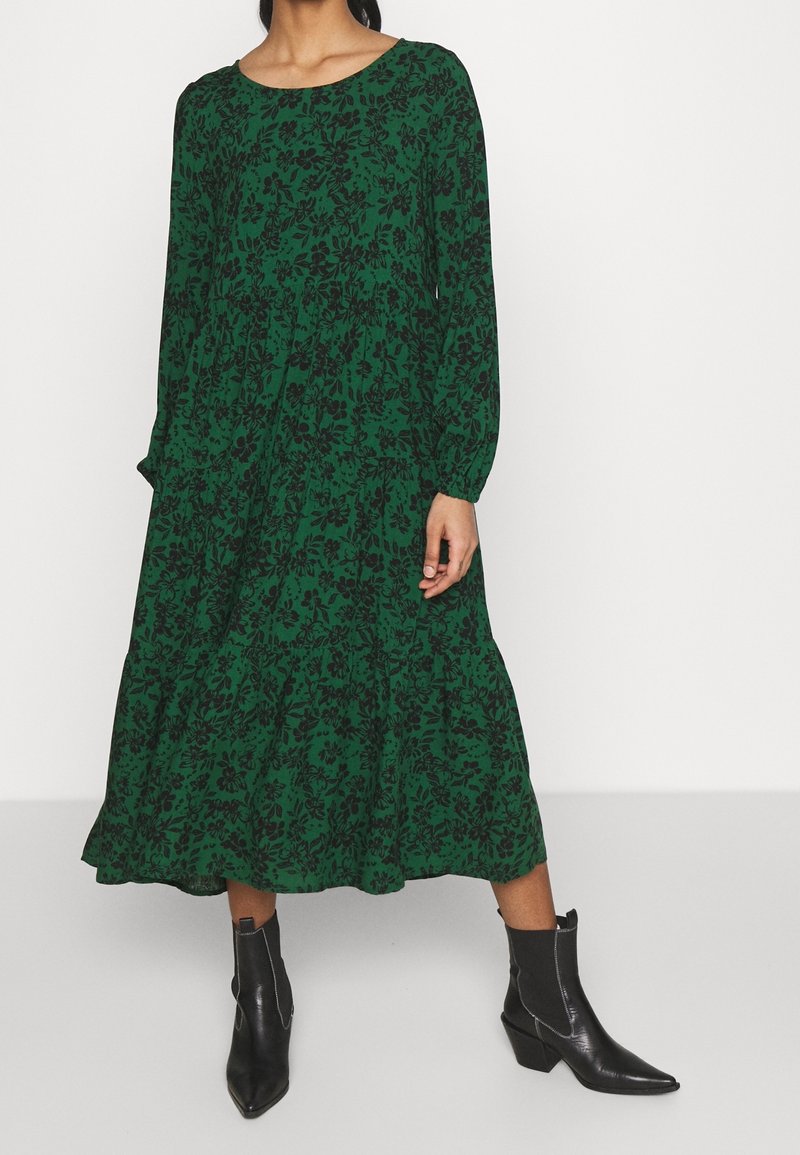 Femme portant une robe verte à manches longues avec imprimé floral noir et des bottines noires à bout pointu, debout devant un fond blanc.