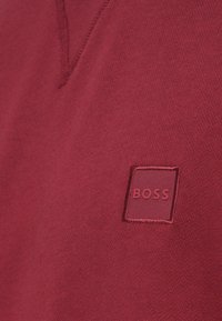 Bordó poló puha anyagból, V-nyakkivágással. Közepén "BOSS" felirattal ellátott hímzett négyzet alakú logóval.