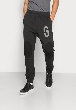 Czarne spodnie dresowe z szarym logo, elastycznym pasem i łatami na kolanach. Dopasowane do czarnych sneakersów z białą podeszwą.