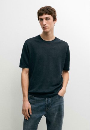 Jonge man met kort bruin haar, gekleed in een donker kortharig vest en blauwe spijkerbroek, staand tegen een effen lichte achtergrond.