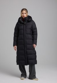 Lange zwarte pufferjas met een capuchon, voorzien van gestikte panelen, een ritssluiting aan de voorkant en zijzakken. Draagt over losse grijze broek en sneakers.
