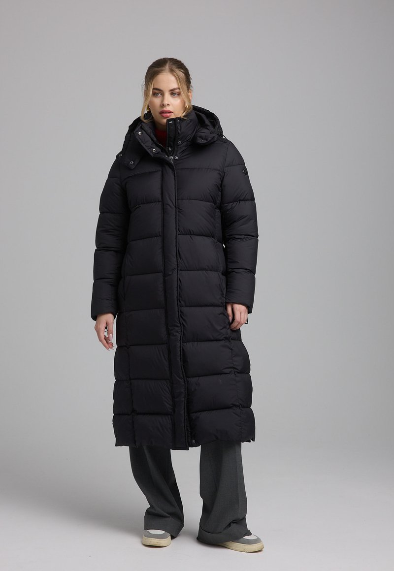 Lange zwarte pufferjas met een capuchon, voorzien van gestikte panelen, een ritssluiting aan de voorkant en zijzakken. Draagt over losse grijze broek en sneakers.