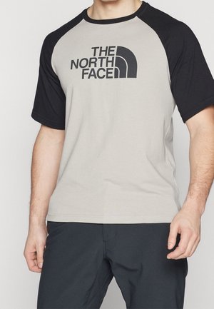 Mann trägt hellgraues und schwarzes Raglan-T-Shirt mit "THE NORTH FACE" Logo auf der Brust, kombiniert mit schwarzer Hose.