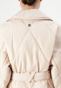 Bakside av en person som har på seg en lys beige quiltet jakke med belte og liten metalllogo nær kragen, håret oppsatt.