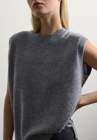 Jeune personne portant un gilet en tricot gris sans manche, posant avec une main sur la hanche devant un fond clair.