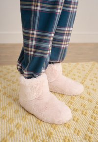 Růžové pantofle s chlupatou podšívkou, zaoblenou špičkou a měkkou texturou, doplněné modro-červenými kostkovanými kalhotami na texturovaném žlutém koberci.