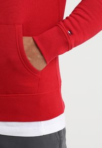 Sudadera roja con capucha, con un bolsillo tipo canguro en el frente, puños y dobladillo acanalados. La tela parece suave con un acabado texturizado. Detalle de logotipo pequeño.