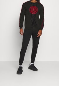 Uomo che indossa un completino sportivo nero con dettagli rossi e logo sulla felpa, scarpe da ginnastica nere e calze bianche, in piedi di fronte a uno sfondo grigio chiaro.