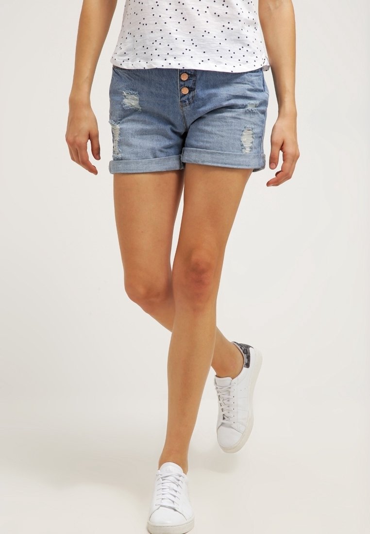 Shorts de mezclilla en azul claro con dobladillo deshilachado, detalles desgastados y cierre de tres botones. Combinados con una blusa blanca con lunares y tenis blancos.