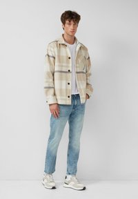 Cremfarbene karierte Jacke mit großen braunen Knöpfen, mit blauen und grauen Streifen, übereinander getragen über einem weißen T-Shirt, kombiniert mit hellblauen Jeans und Sneakers.