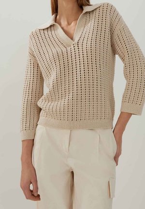 Kvinde iført en beige åbenstrikket sweater med krave og trekvartærmer kombineret med lysebeige højttaljede bukser.
