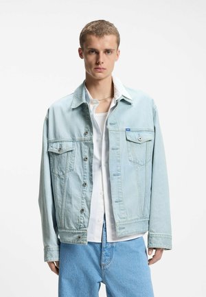 Młody mężczyzna ubrany w jasnoniebieską kurtkę jeansową, białą koszulę, biały podkoszulek i jasnoniebieskie dżinsy, stojący na tle gładkiej białej ściany.