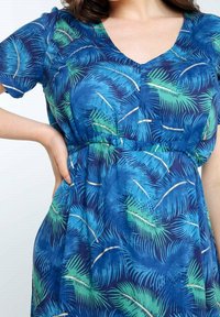 Femme portant une robe bleue avec des motifs de feuilles tropicales vertes et bleu marine et un décolleté en V, main reposant sur la hanche.