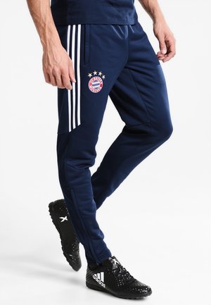 Person trägt marineblaue FC Bayern München Trainingshose mit weißen Streifen und schwarze Adidas Fußballschuhe, steht vor weißem Hintergrund.
