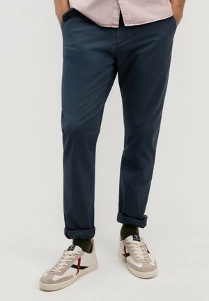 Hombre con pantalones azules oscuros remangados, calcetines verde oliva y zapatillas blancas con detalles en rojo y negro sobre un fondo neutro.