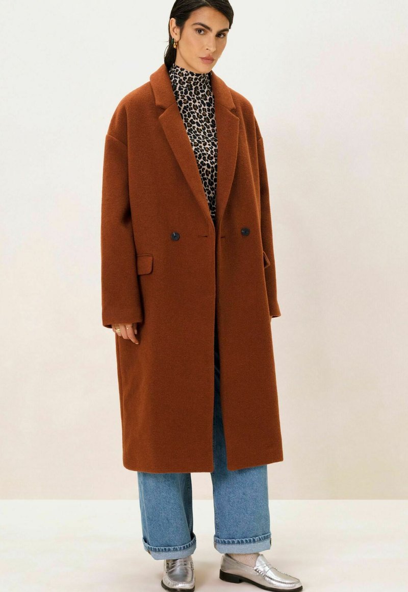 Manteau marron surdimensionné à double boutonnage avec revers, porté sur un haut à imprimé léopard noir et blanc, associé à un jean large bleu clair et des chaussures argentées.