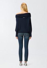 Mornarsko plavi pulover bez ramena s rebrastim izrezom i manžetama, u kombinaciji s uskim trapericama i srebrnim visoko potpeticama.