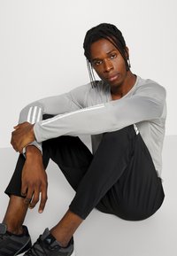 Camiseta atlética de manga larga gris con rayas blancas en las mangas, combinada con pantalones negros ajustados y zapatillas negras, en posición sentada.