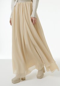 Jupe maxi fluide beige en tissu léger, dotée d'une taille froncée et d'une texture douce et transparente, associée à des bottes à talons bloc légers.