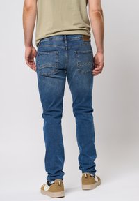 Jeans de mezclilla azul con un corte slim, que cuentan con bolsillos traseros y detalles de costura sutiles. Combinados con zapatillas color marrón claro y una camisa beige.