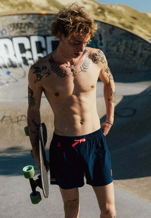 Jeune homme torse nu avec des tatouages et des cheveux bouclés tenant un skateboard dans un skatepark ensoleillé avec des graffitis sur la rampe.