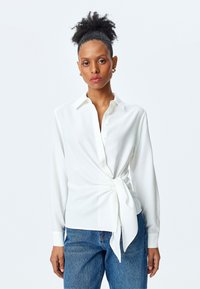 Camisa blanca con mangas largas, cuello y detalle de lazo en la parte frontal. Fabricada en tejido suave. Combinada con vaqueros de tela vaquera azul.