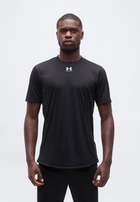 Νεαρός άνδρας στέκεται με το σώμα στραμμένο προς τα εμπρός φορώντας ένα μαύρο αθλητικό μπλουζάκι με κοντά μανίκια Under Armour, σε ένα απλό ανοικτό φόντο.