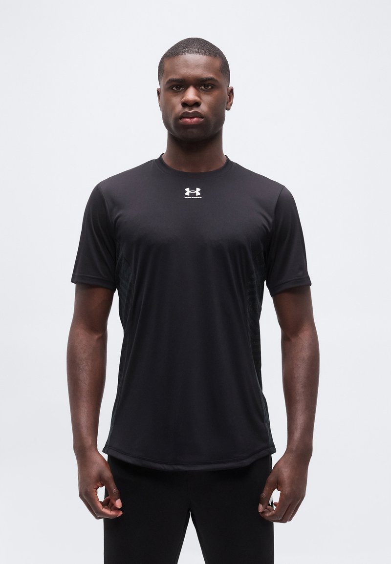 Νεαρός άνδρας στέκεται με το σώμα στραμμένο προς τα εμπρός φορώντας ένα μαύρο αθλητικό μπλουζάκι με κοντά μανίκια Under Armour, σε ένα απλό ανοικτό φόντο.