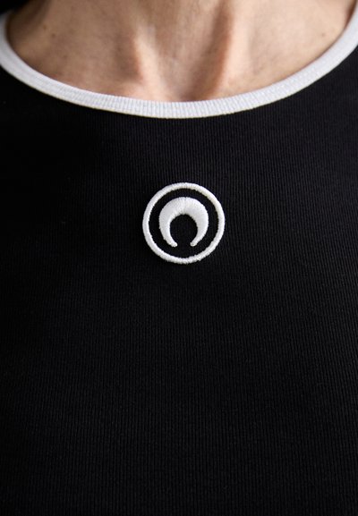 Črna rebrasta majica z izrezom, ki ima bel krožni vezen emblen na vratu, z kontrastnim belim rebrastim ovratnikom.