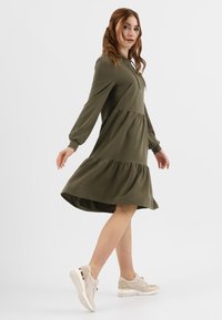 Abito lungo a maniche lunghe verde oliva con gonna a strati, dotato di cappuccio e lacci. Modello che indossa sneaker beige con accenti metallici.