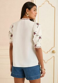 White blouse with floral embroidery at puff sleeves and scalloped trim; paired with blue denim shorts; smooth texture. Gold hoop earrings visible.