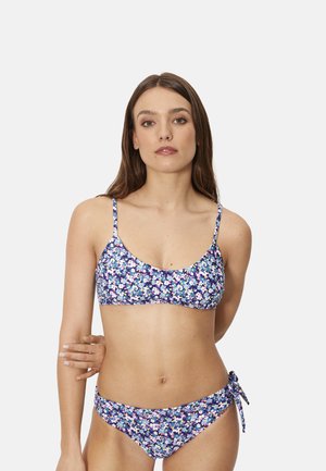 Mujer con un bikini floral azul de tirantes finos y braguitas con lazo lateral, de pie frente a un fondo gris claro liso.