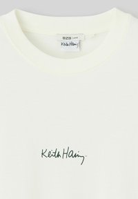 T-shirt ample couleur crème en coton avec un col rond. Il présente un logo imprimé vert foncé avec l'inscription "Keith Haring" près de l'ourlet.