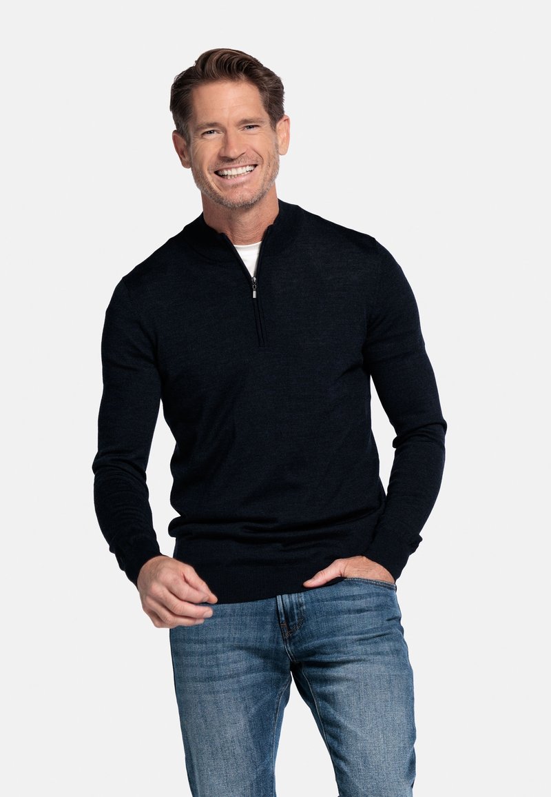 Donkerblauwe pullovertrui gemaakt van zacht materiaal, met een kwart-rits kraag, geribbelde manchetten en een aansluitend design, gedragen over een wit shirt.