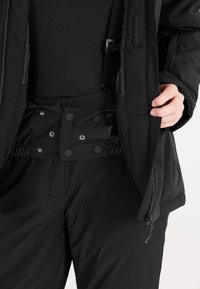 Schwarz isolierte Jacke mit Reißverschluss, verstellbaren Bündchen und Druckknöpfen. Getragen über schwarzen Hosen mit elastischem Bund und seitlichen Reißverschlusstaschen.