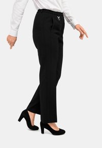 Schwarze maßgeschneiderte Hose mit elastischem Bund, seitlichen Taschen und eleganten Details, kombiniert mit schwarzen Wildleder-High Heels.