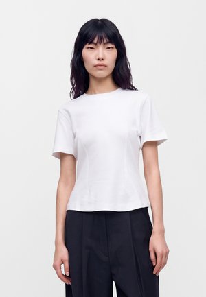 Jeune femme aux longs cheveux foncés portant un t-shirt blanc uni à manches courtes et un pantalon noir, faisant face à l’avant sur un fond blanc.