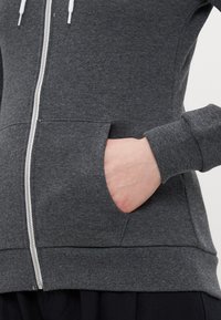 Sudadera con cremallera de color gris oscuro, fabricada en tela suave, con un bolsillo frontal y detalles de puños de canalé. Cremallera plateada y cordones blancos.