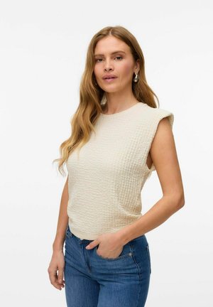 Cremefarvet, tekstureret, ærmeløs top med rund halsudskæring, der viser et subtile vaffelmønster, parret med blå, højtaljede jeans.
