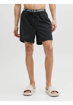 Mężczyzna ubrany w czarne kąpielówki Jack & Jones ze sznurkiem oraz beżowe sandały wsuwane, stojący na jednolitym jasnym tle.