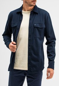 Dunkelblaues Hemd mit Knopfverschluss, zwei Brusttaschen und Kragen, aus einem strukturierten Stoff, über einem hellbeigen T-Shirt getragen.