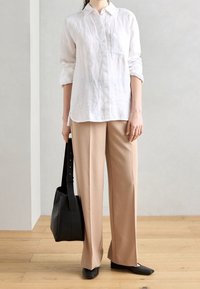 Witte linnen button-up shirt met lange mouwen en zak, gecombineerd met beige wijde broek, zwarte platte schoenen en een zwarte tote bag.