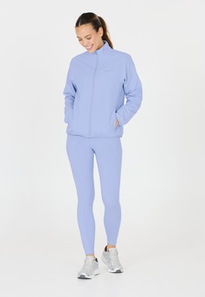Ljuslila träningsjacka med dragkedja fram, sidofickor och matchande leggings. Modellen bär silverfärgade sneakers. Slät textur genom hela plagget.