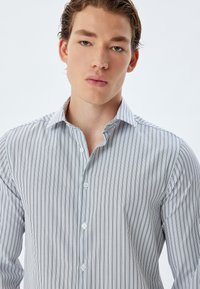 Camisa blanca de botones con finas rayas verticales negras, un cuello clásico y mangas largas con puños estructurados.