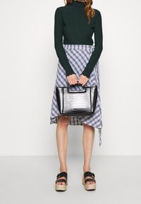Svart krokodilprickig handväska med strukturerad design och topphandtag. Modellen bär en grön ribbad tröja och en blå rutmönstrad omslagsskjorta.
